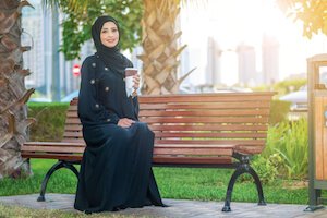 Smiling Arab woman dressed in an abaya sitting on a bench in sunshine - image by shutterstock.com